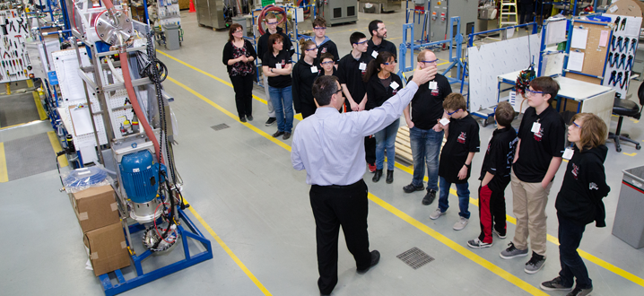 a businessman in blue shirt and slacks is pointing out a feature to a group of students on an industry tour